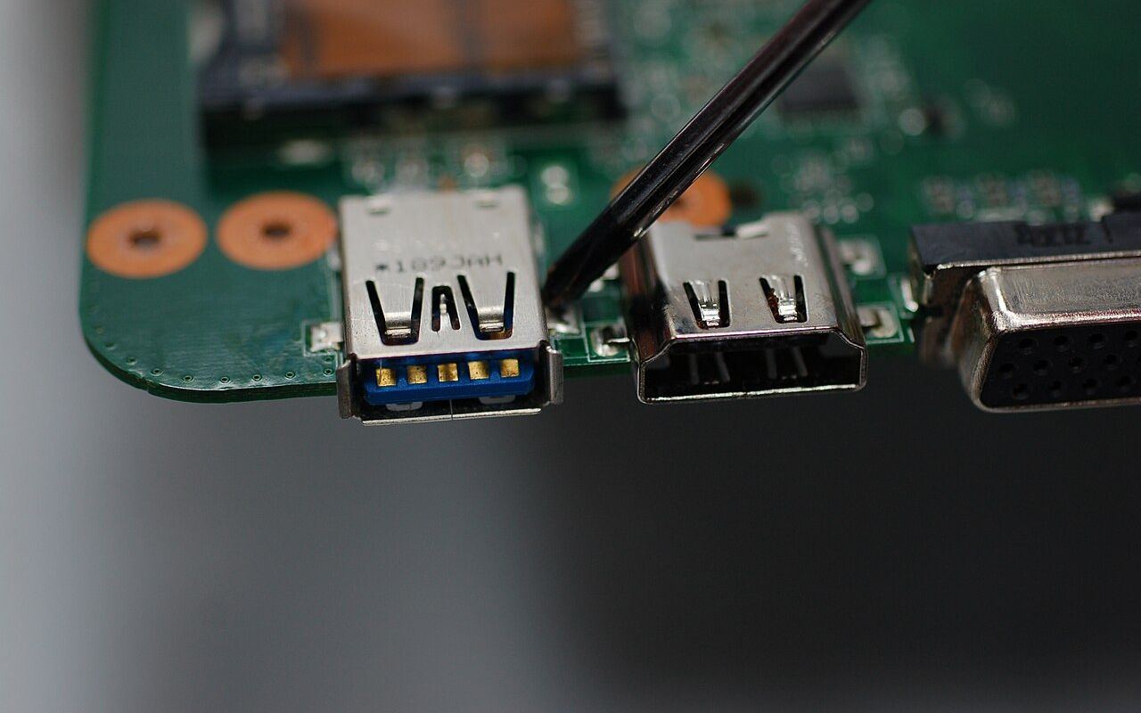 USB port solder points on laptop motherboard (closeup, indicated by screwdriver)
